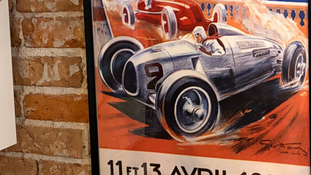 A framed vintage Monaco racing poster is being hung on a red brick wall by a person whose head and arms are visible. The poster features a classic silver race car and the text 'MONACO' and '11 et 13 AVRIL 1936'.