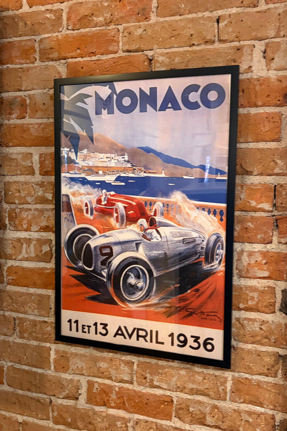 A framed vintage Monaco racing poster hanging on a red brick wall. The poster features a silver race car on an orange track with blue water and mountains in the background, and the text 'MONACO 11th-13th April 1936'.